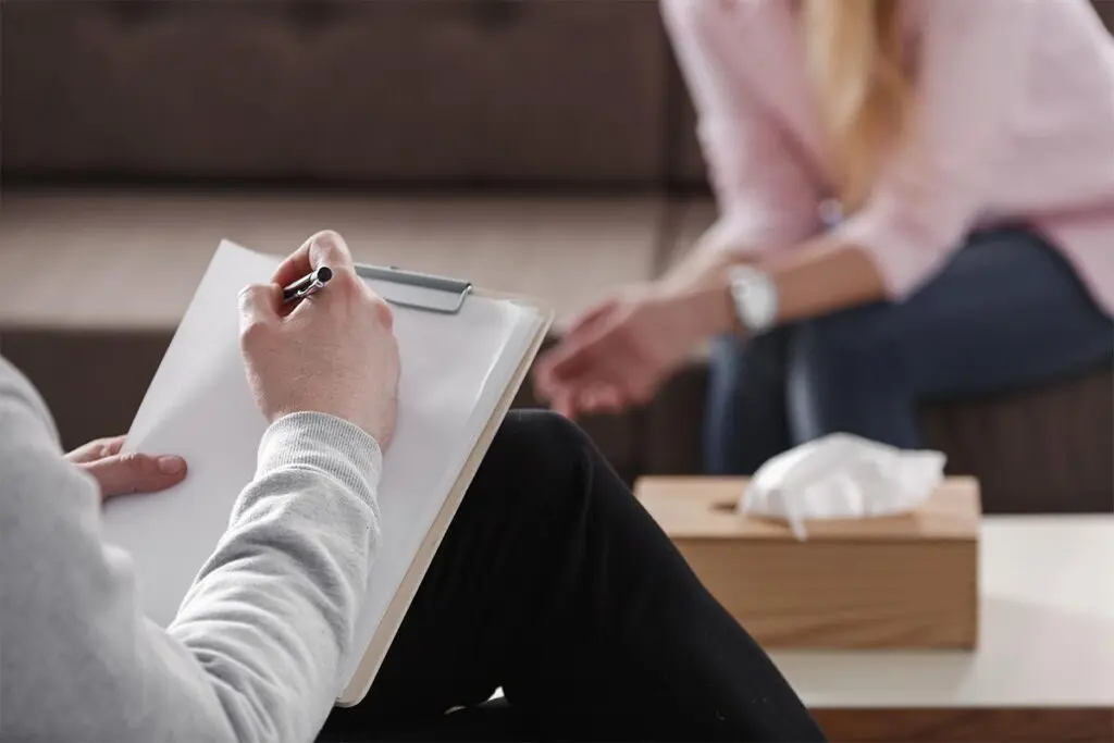 A person with long hair sits on a sofa, partly visible, while another person in the foreground writes on a clipboard. A box of tissues is on a table between them, suggesting a therapy session. The setting is calm and professional.