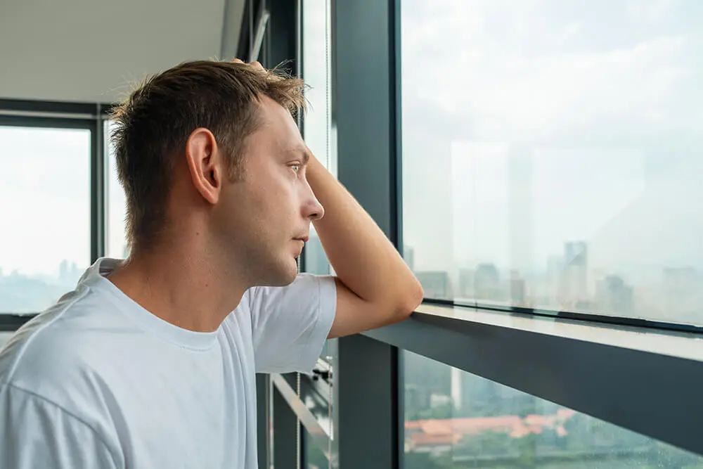Man in a white t-shirt looking thoughtful, with one hand resting on his head, gazing out a large window. The window offers a view of a hazy cityscape with buildings in the distance.