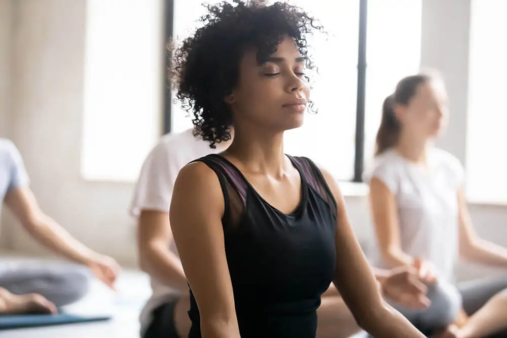 A person with curly hair sits cross-legged in a yoga class, eyes closed, wearing a black sleeveless top. Other participants are blurred in the background, sitting in similar poses. The room is well-lit with large windows.