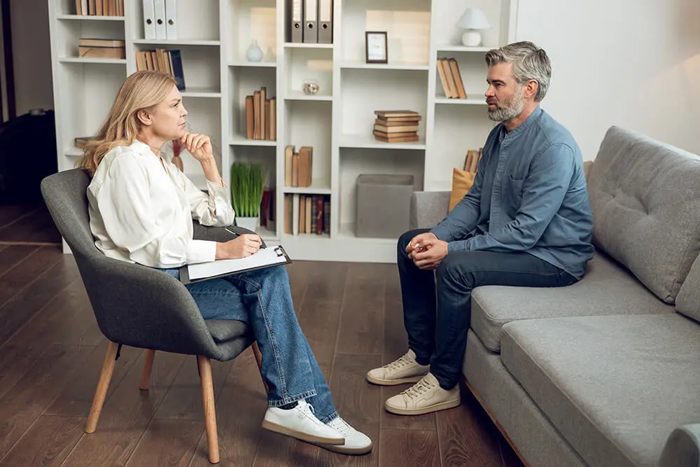 A woman and a man are sitting across from each other in a modern living room. The woman is holding a clipboard, appearing to be taking notes, while the man listens attentively. They are surrounded by contemporary decor and bookshelves.