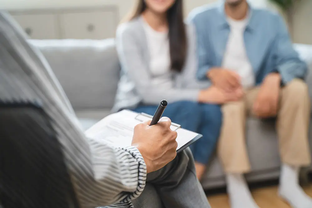 A couple sits on a couch during a counseling session. Theyre holding hands, facing a therapist who is taking notes on a clipboard. The setting is calm and professional, with soft lighting and neutral tones.