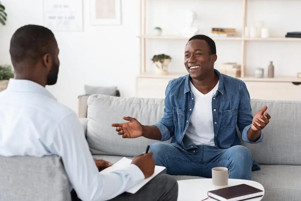 Two men sit on a sofa. One is holding a clipboard and pen, while the other gestures with open hands, smiling. A mug and a book are on the table between them. Shelves with decor are in the background.