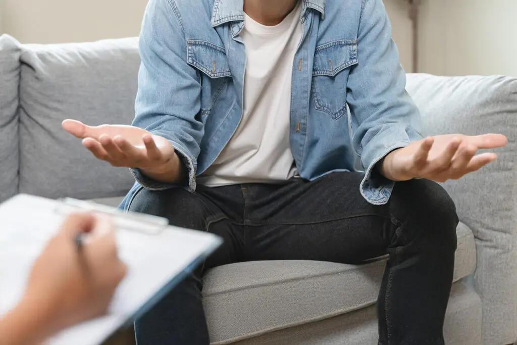 A person in a denim jacket and white t-shirt sits on a couch, gesturing with open hands. Another person holds a clipboard in the foreground, suggesting a conversation or counseling session. The setting is casual and indoors.