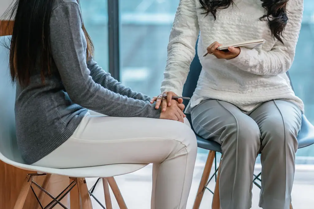 Two women seated, one in a white sweater holding a notebook, gently holding hands with another in a gray sweater. The setting appears to be a supportive or counseling environment, with natural light coming through a window.