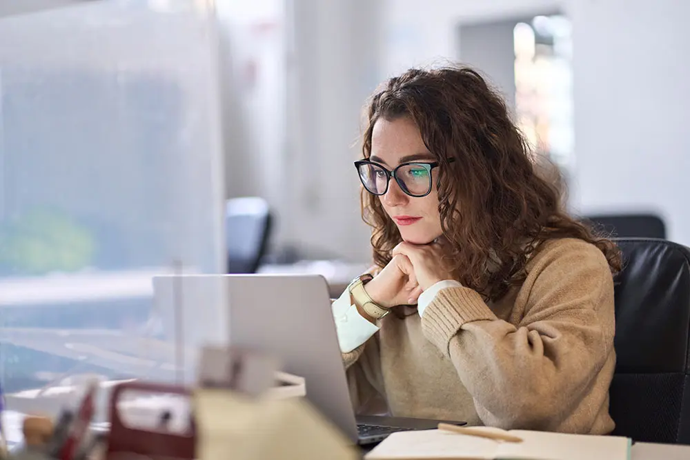 A woman with curly hair and glasses sits at a desk, looking intently at a laptop screen in an office setting. She rests her chin on her hand and appears focused on her work.