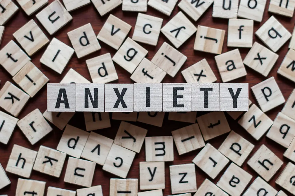 Wooden letter tiles scattered on a table. The word ANXIETY is prominently spelled out in the center using these tiles.