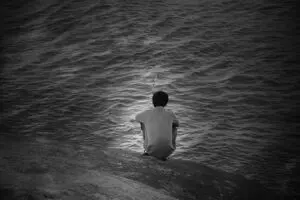 A person sitting at the edge of a rocky surface, fishing in dark, rippling water. The scene is captured in black and white, creating a serene and contemplative atmosphere.
